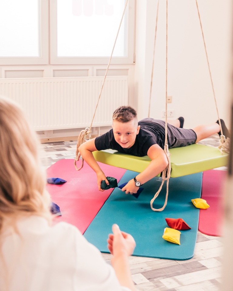 Junge liegt bäuchlings auf einer Plattformschaukel im Therapieraum